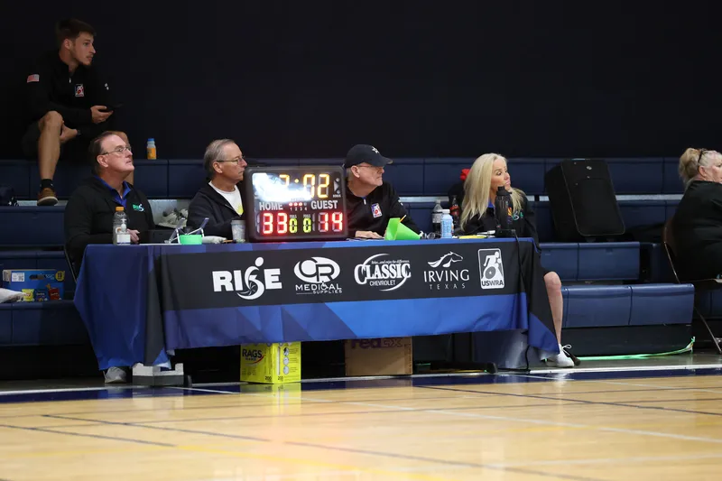 Volunteers staff the scorer's table under the scoreboard and RISE banners