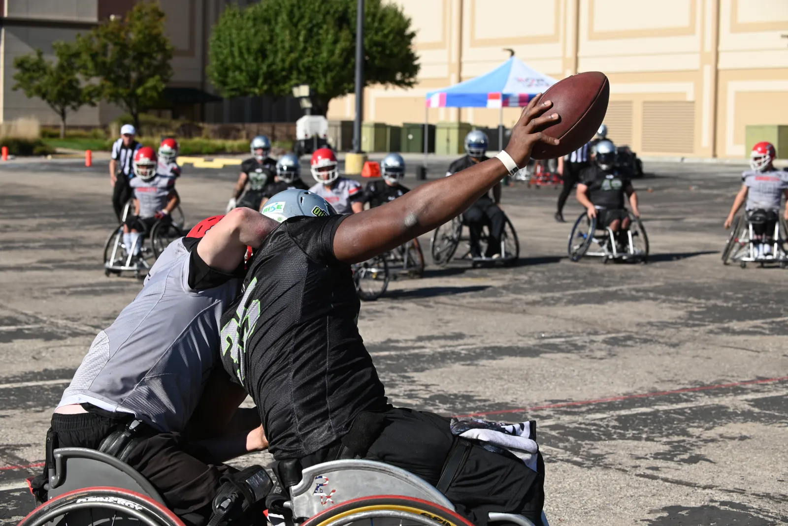 An Outlaws player in a black jersey extends one arm to hold the football out of reach as a gray-jersey, red-helmet defender leans hard into his shoulder on the sunlit lot.