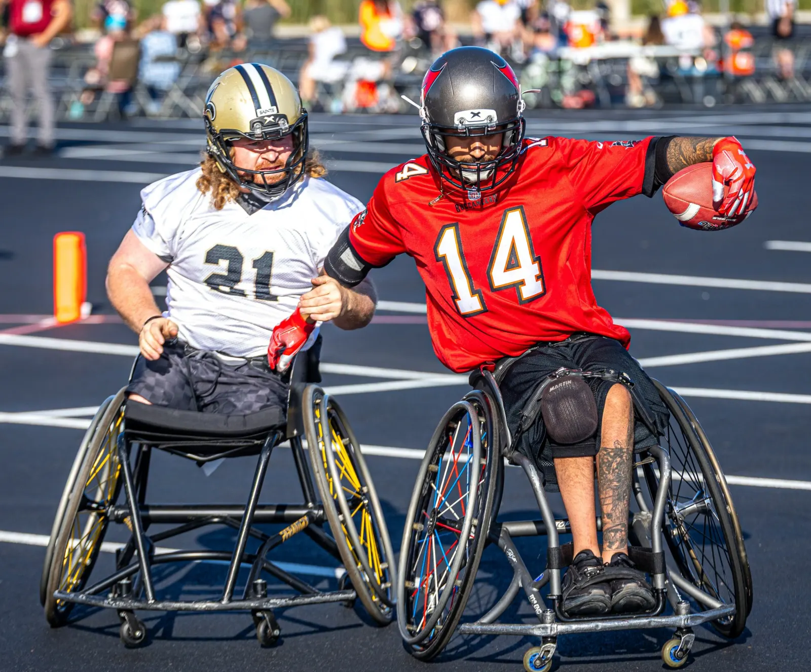 A Buccaneers player in a red number 14 jersey holds the ball out to the side while a Saints defender reaches in.