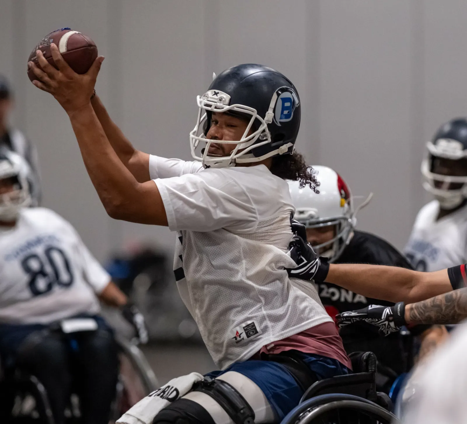 A player in a white jersey and navy helmet cocks the football back to throw amid a cluster of opponents.
