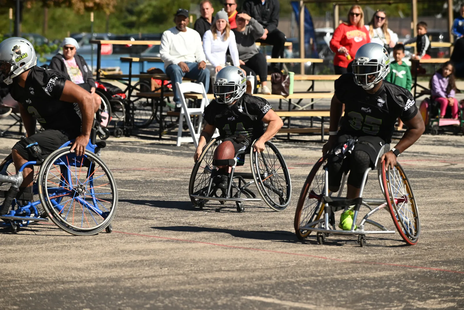 An Irving Outlaws player in a black jersey and silver helmet drives forward with the football resting in his lap, flanked by two silver-helmet teammates as spectators watch from benches.