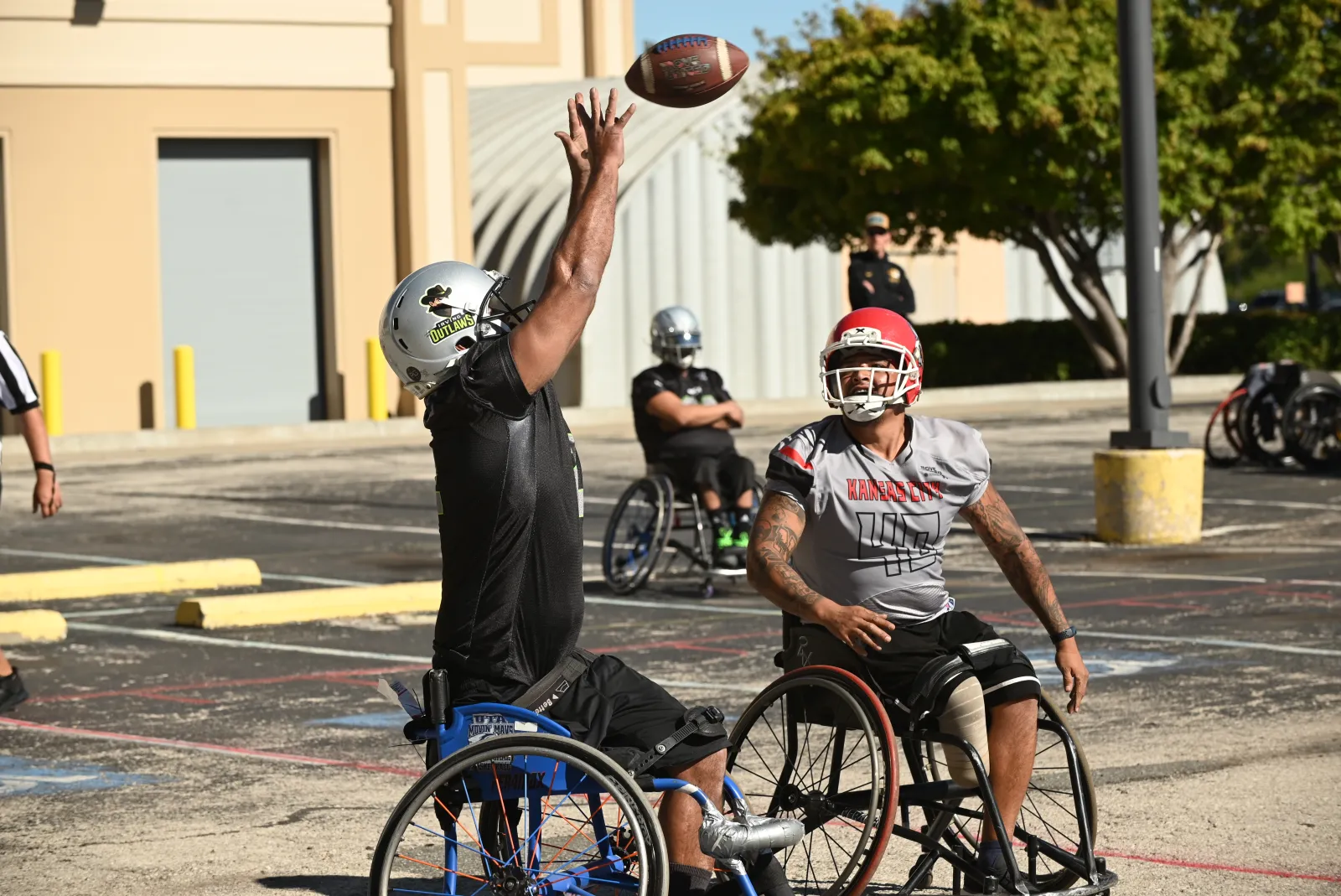 An Irving Outlaws player in black stretches one arm high to pull in a spiraling football while a gray Kansas City number 46 player in a red helmet looks on.
