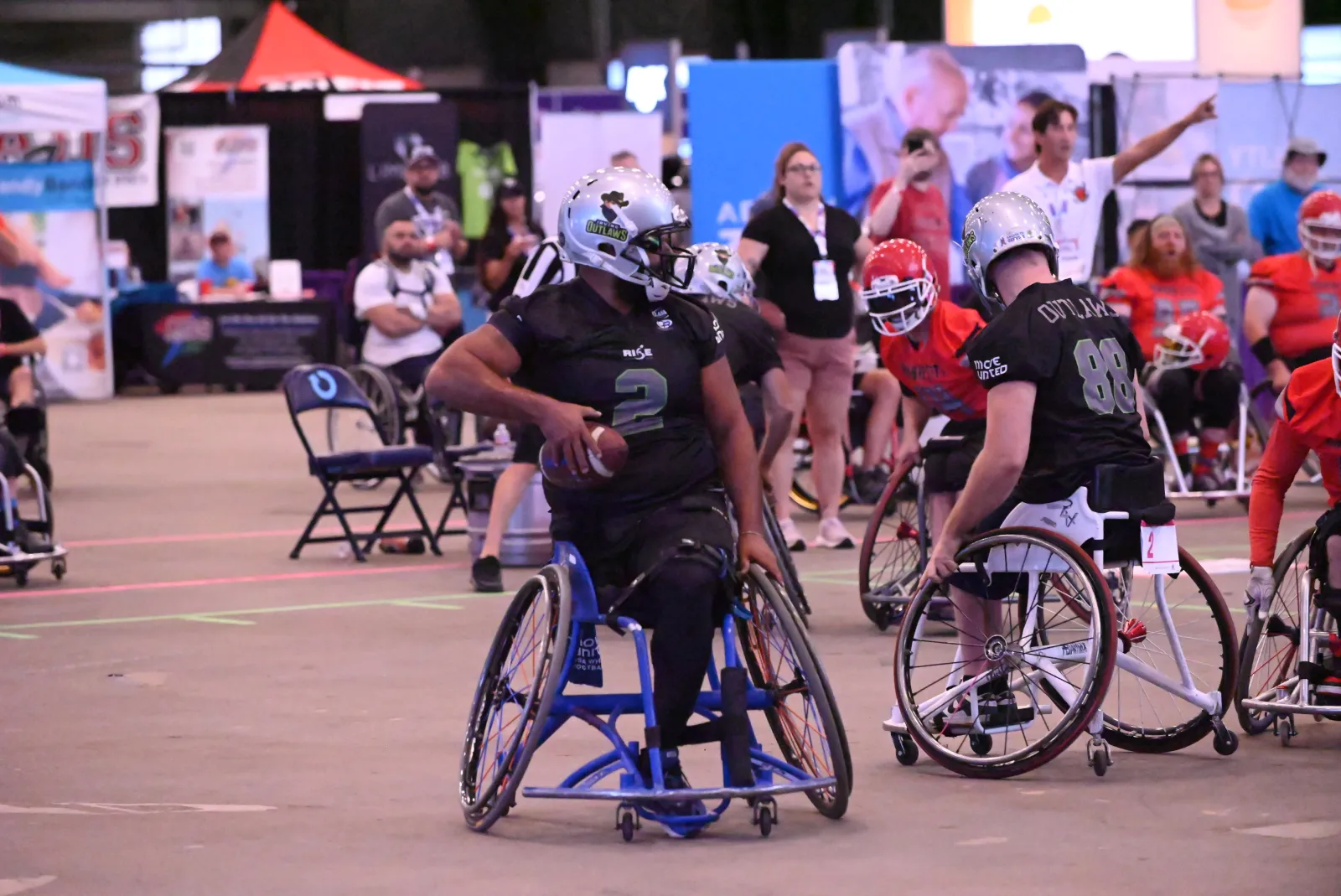 An Irving Outlaws player in a black number 2 jersey and silver Outlaws helmet cradles the football at an indoor arena expo, with teammate number 88 and red-jersey opponents nearby.