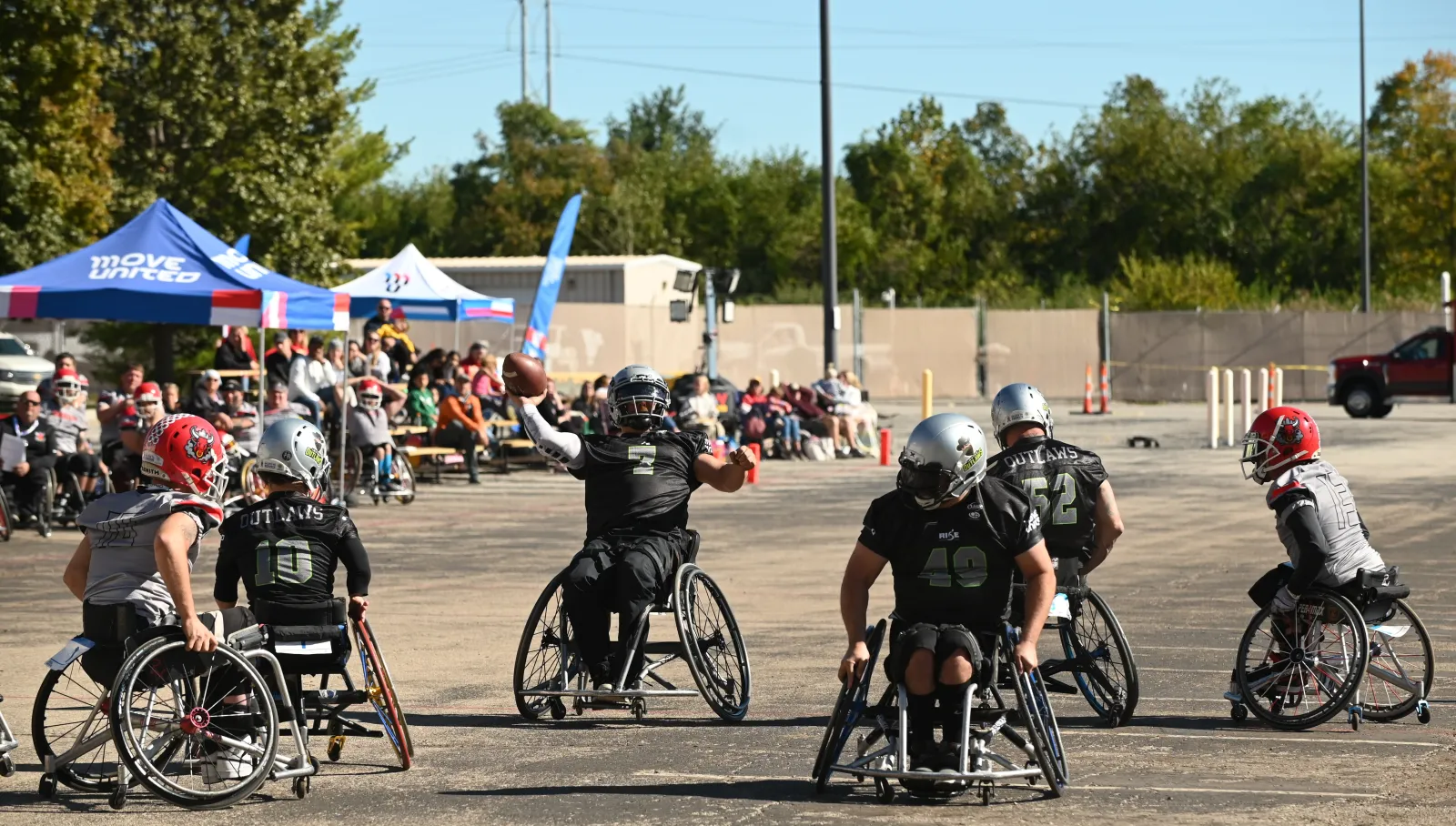 An Irving Outlaws quarterback in a black number 7 jersey cocks the football back to throw while silver-helmet teammates and red-helmet defenders spread across the paved lot around him.