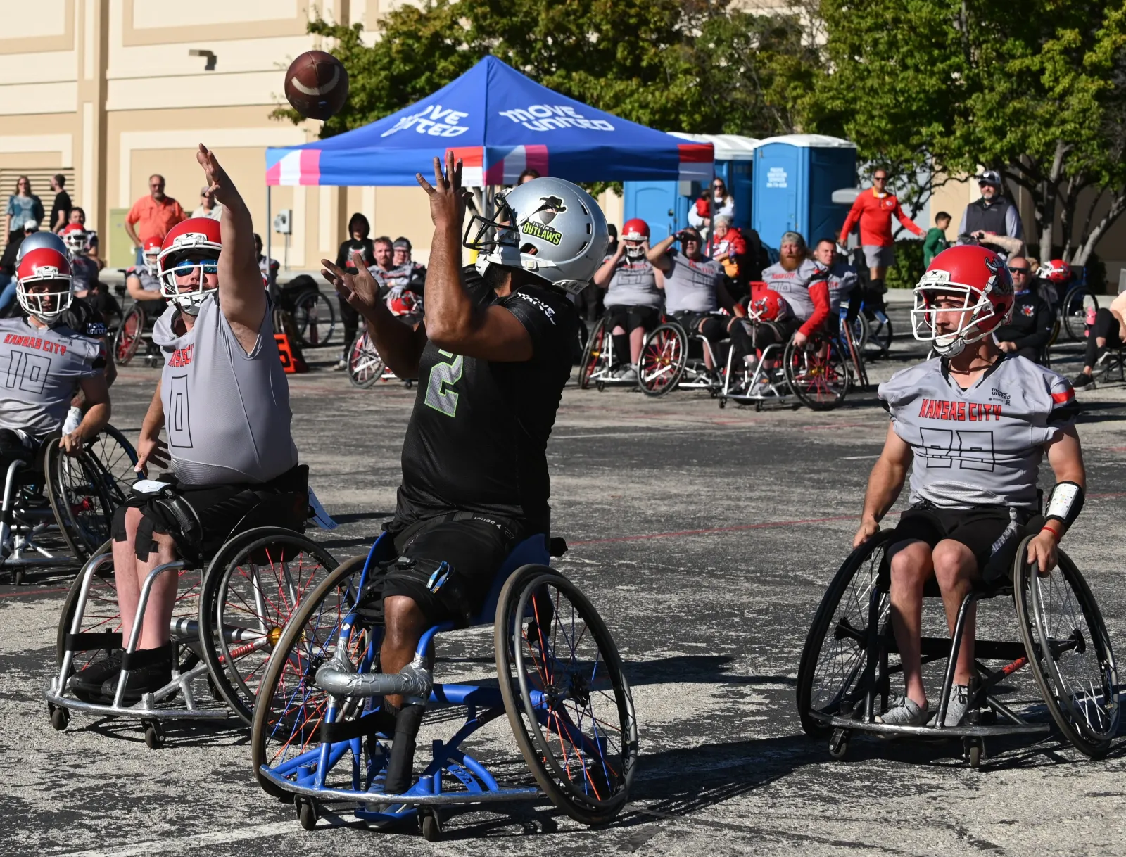 An Irving Outlaws player in a black number 2 jersey and silver helmet reaches both hands up for a football dropping out of the air, flanked by gray-jersey Kansas City players in red helmets.