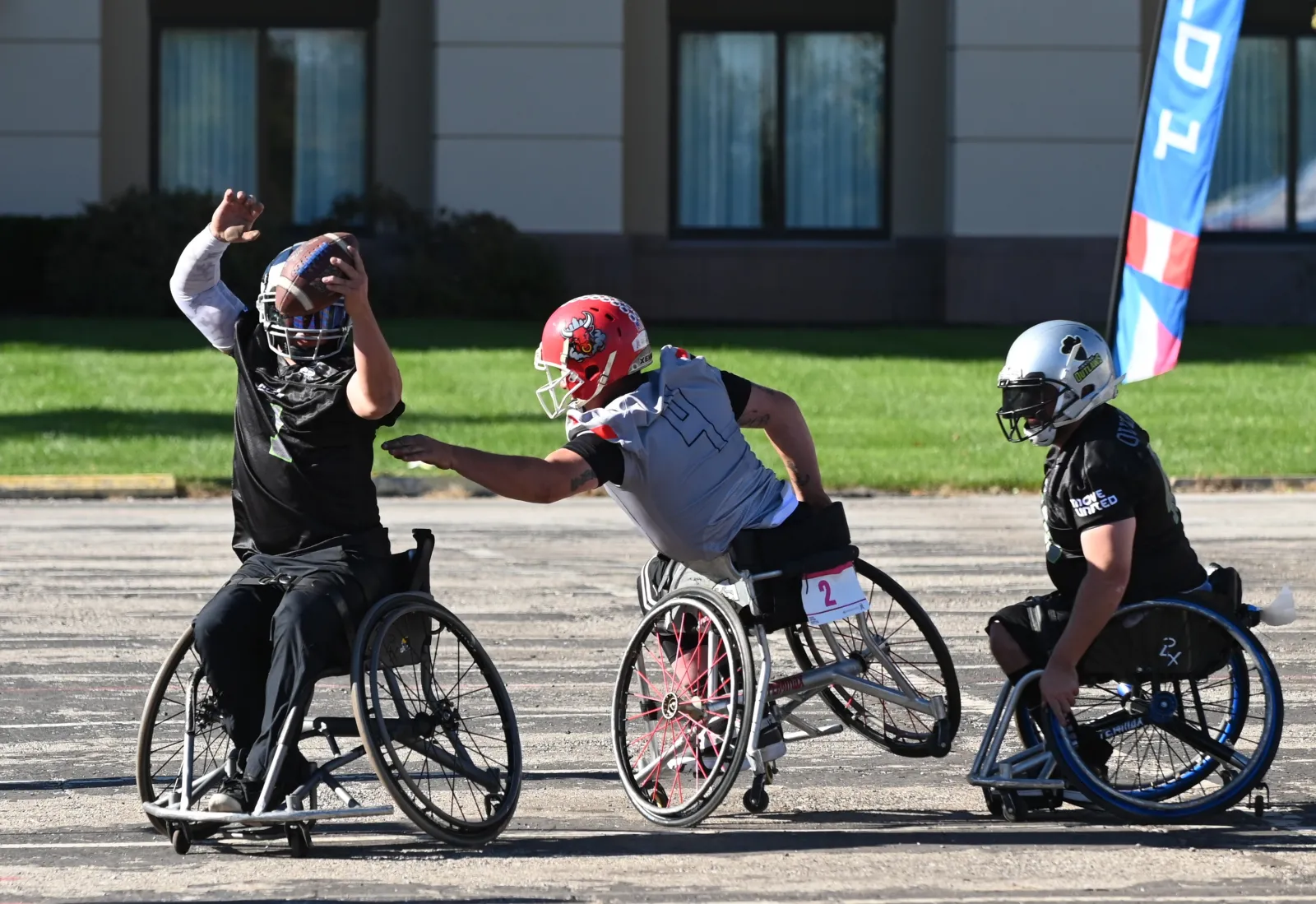 A player in a black jersey raises the football overhead to pass while a gray-jersey opponent in a red bull-logo helmet reaches across to block and a silver-helmet Outlaws player closes in.
