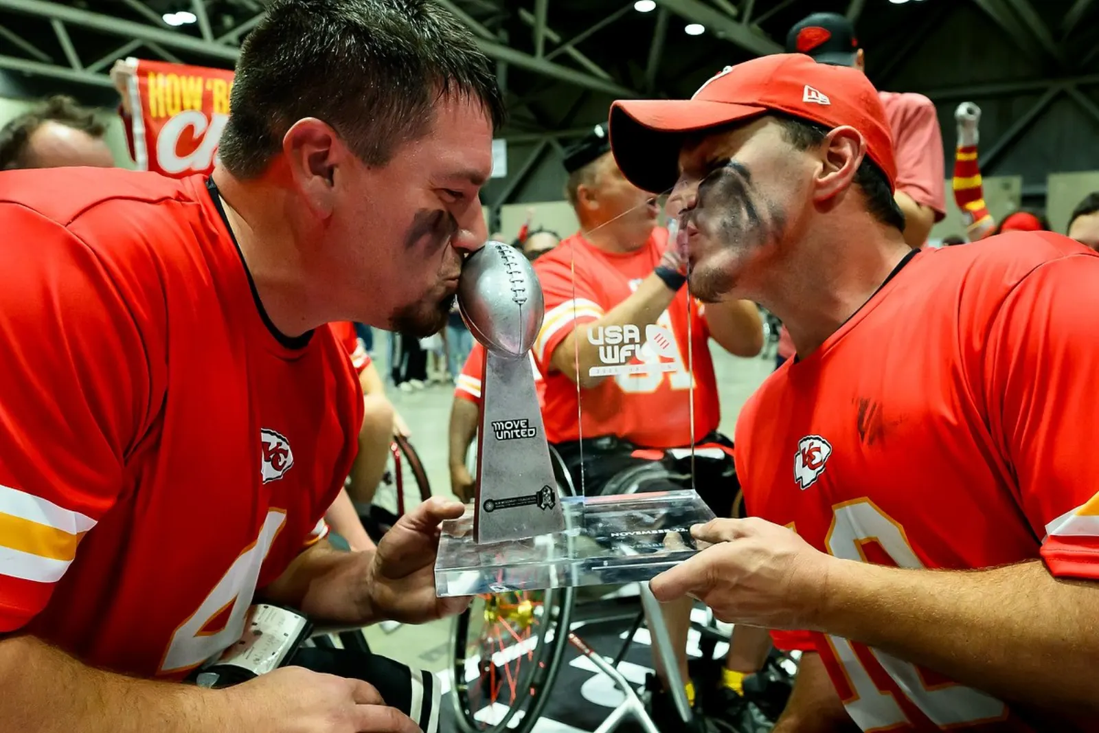 Two Kansas City players in red jerseys lean in to kiss the silver championship trophy held between them.