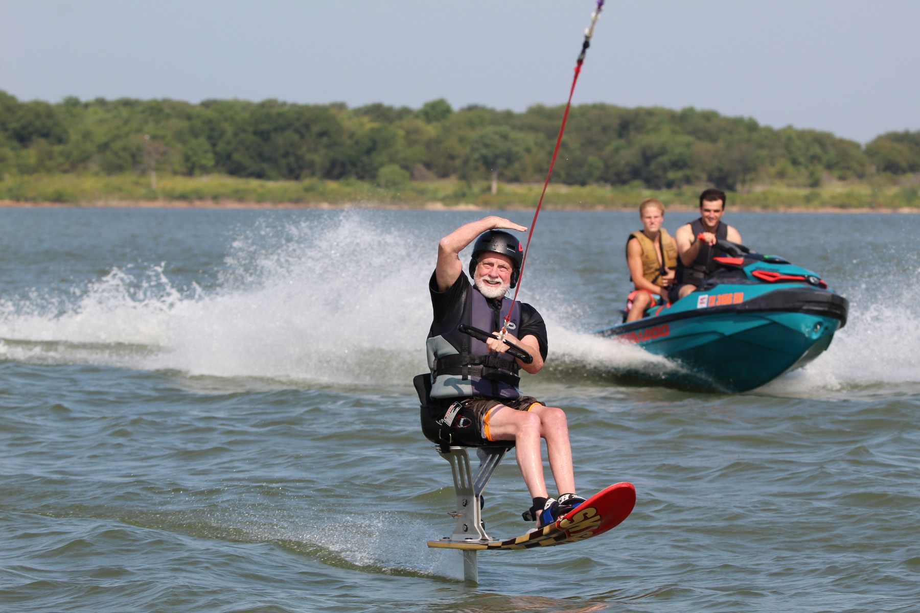 An athlete smiles while riding a seated water ski, with a personal watercraft behind.