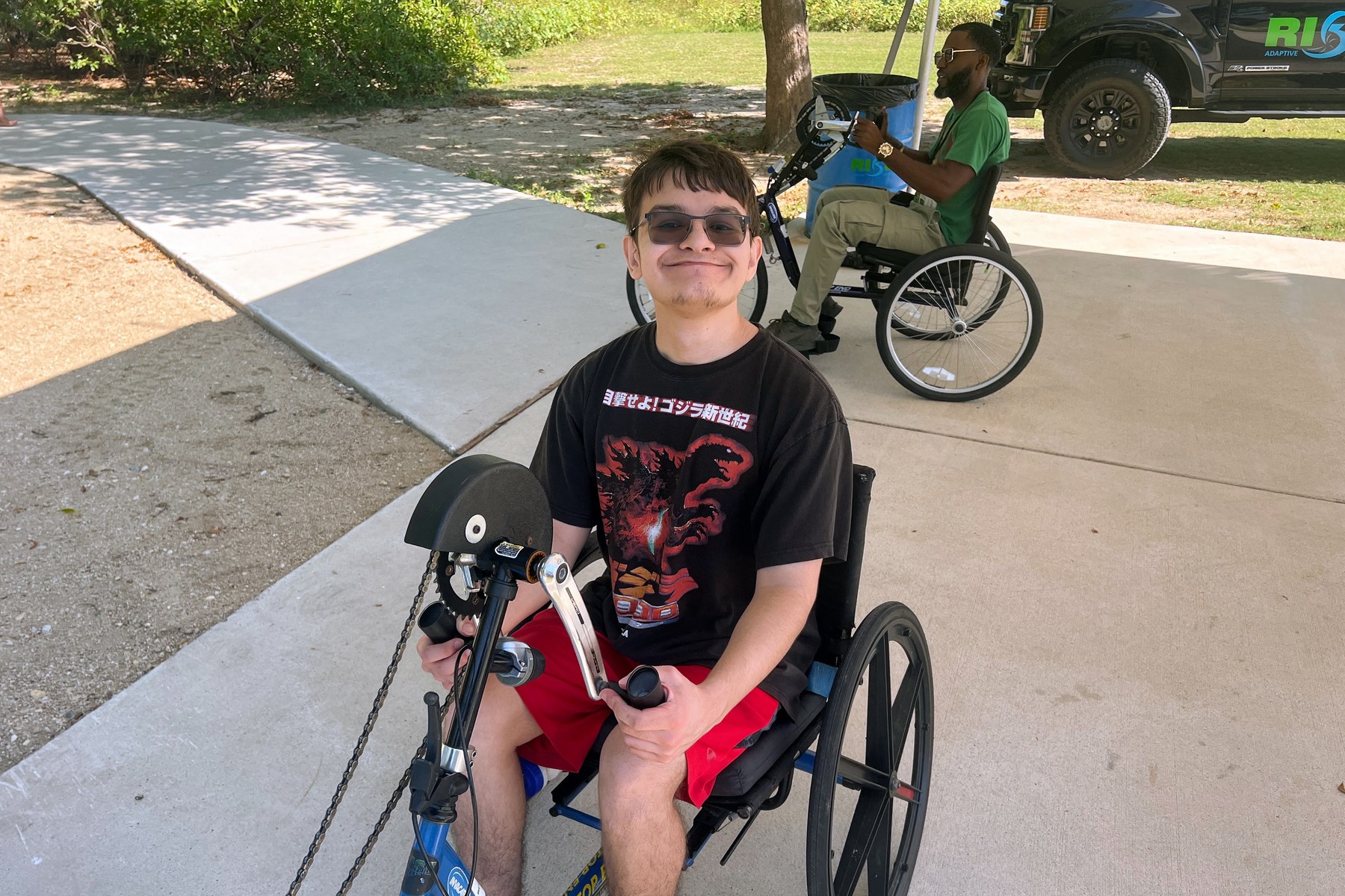 Two people use handcycles on a shaded park path beside a RISE vehicle.
