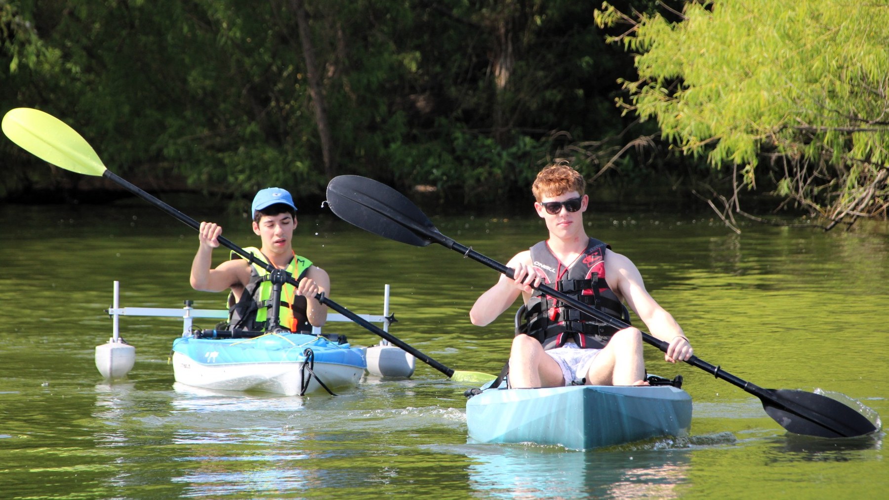 Two people in life jackets paddle blue kayaks beside a wooded shoreline.