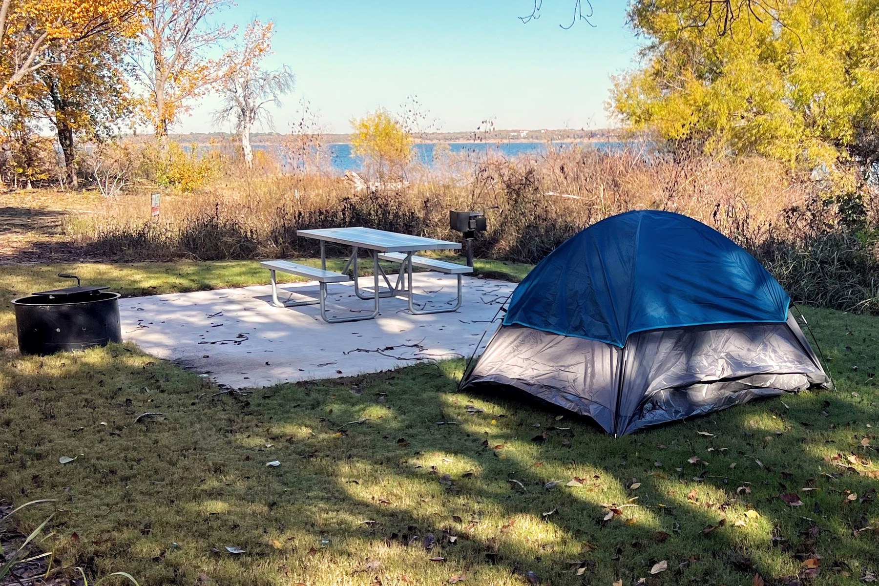 A blue tent stands beside a concrete campsite pad, picnic table, and fire ring overlooking Lake Grapevine.
