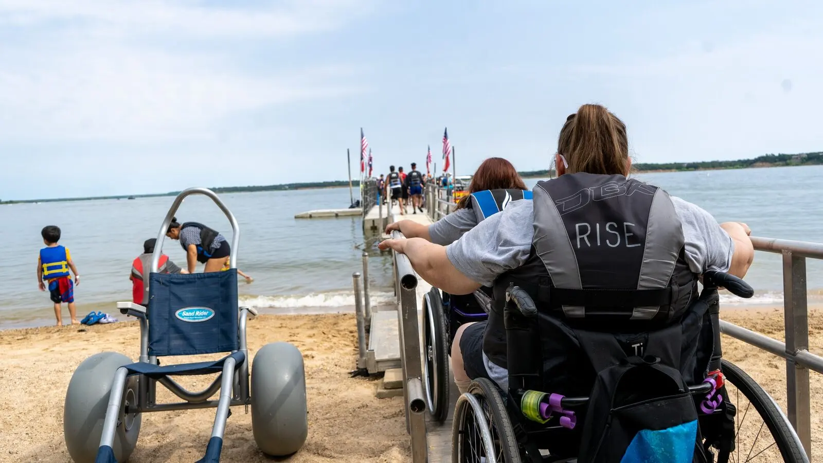 Two wheelchair users, one in a RISE life vest, roll up a ramp toward the Lake Grapevine dock past a beach wheelchair on the sand.