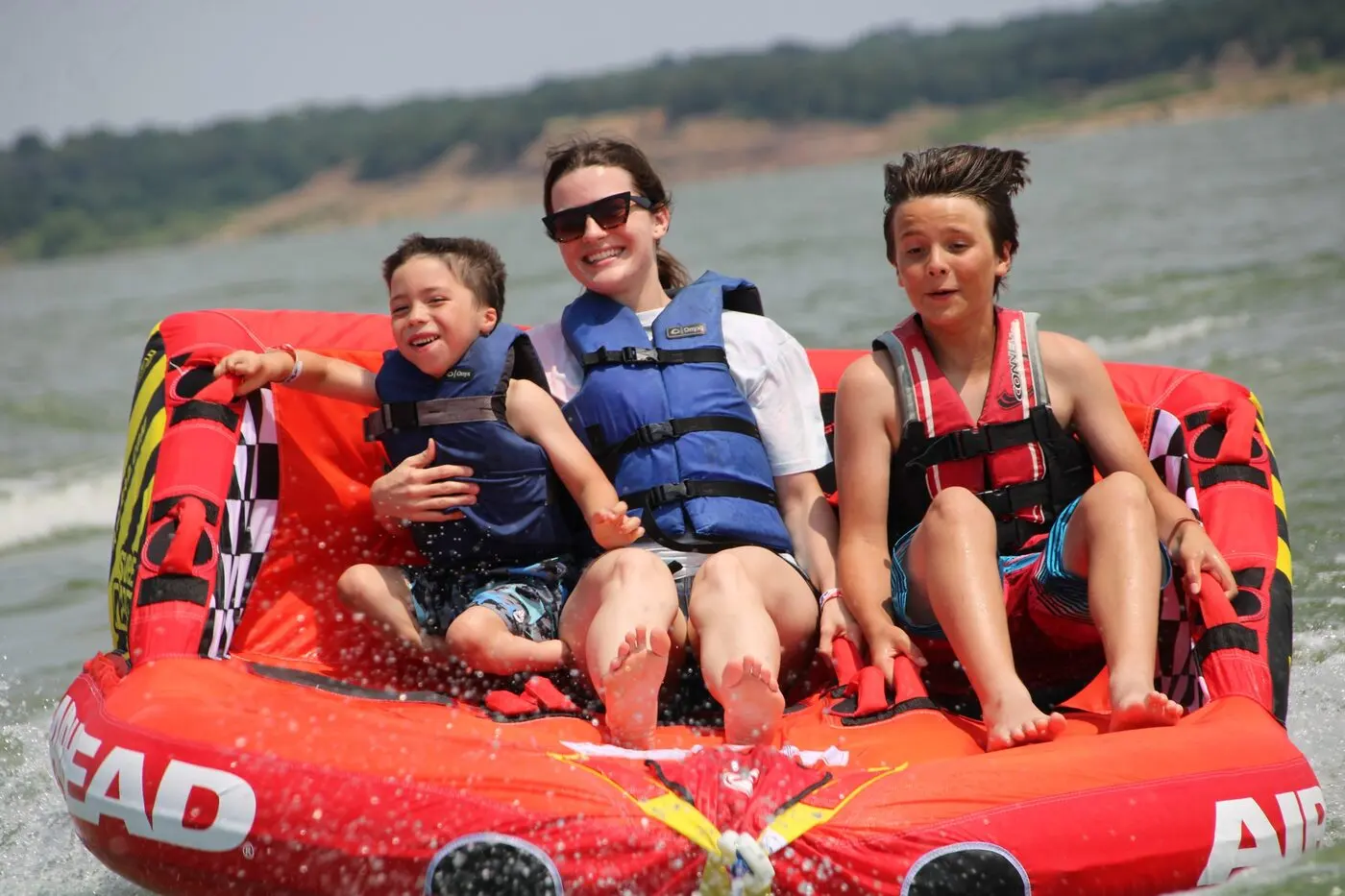 A woman and two boys in life jackets grin while riding a red inflatable tube across Lake Grapevine.