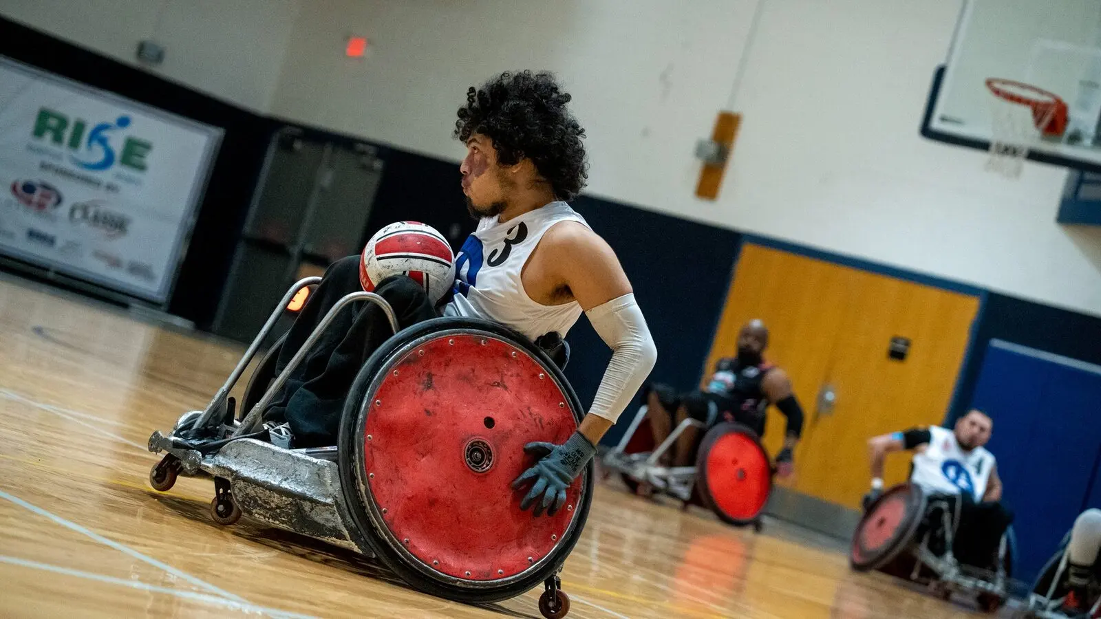 A wheelchair rugby player in a white number 3 jersey cradles the ball on his lap during a gym match, with other players and a basketball hoop behind.