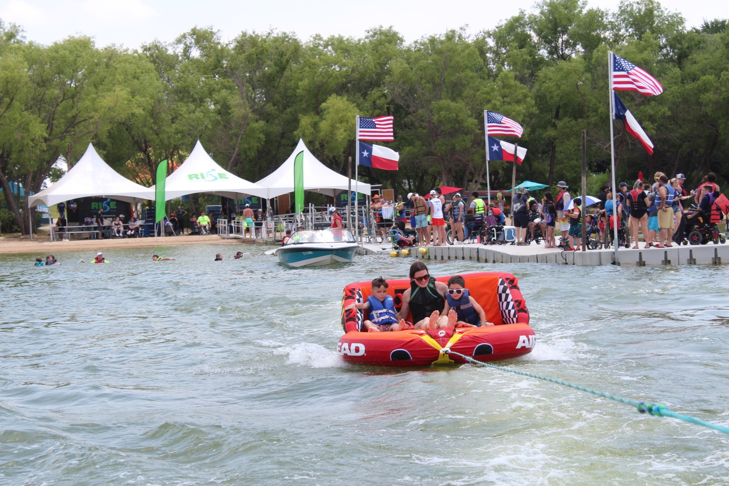 A woman and two boys in life jackets ride a towed tube on Lake Grapevine as RISE tents, US and Texas flags, and wheelchair users line the dock behind.