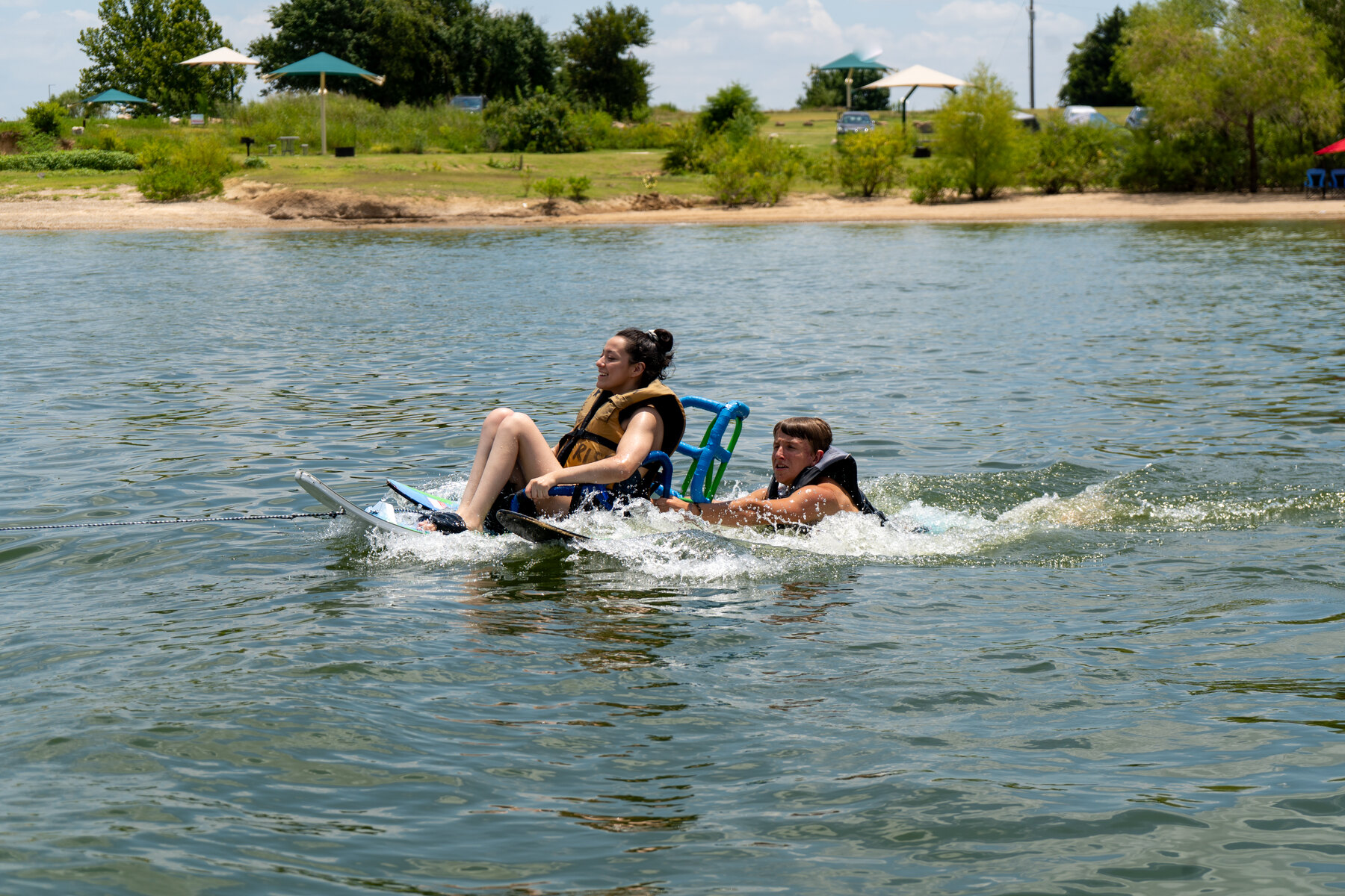 A support person swims beside an adaptive athlete riding a seated water ski near shore.