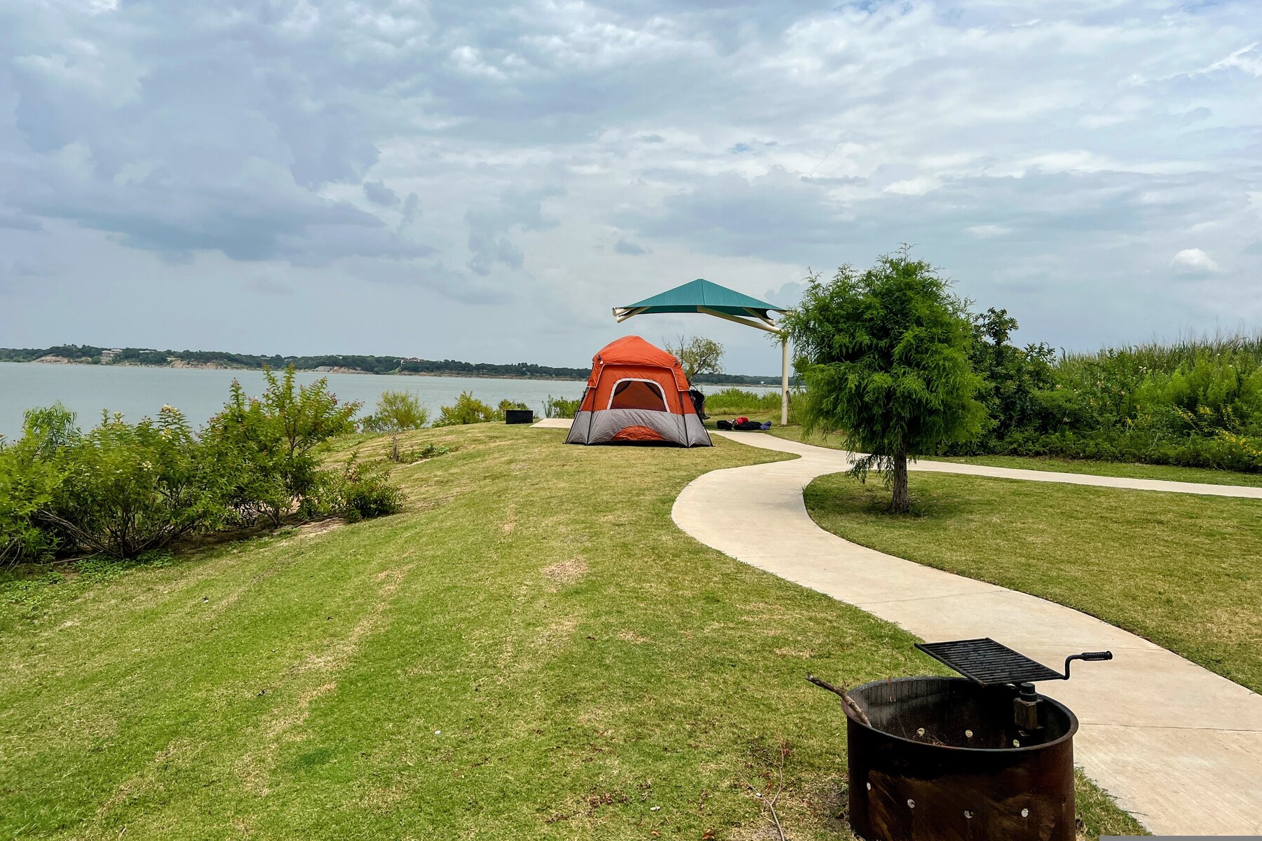 An orange tent stands beside a concrete campsite path and fire ring overlooking Lake Grapevine.